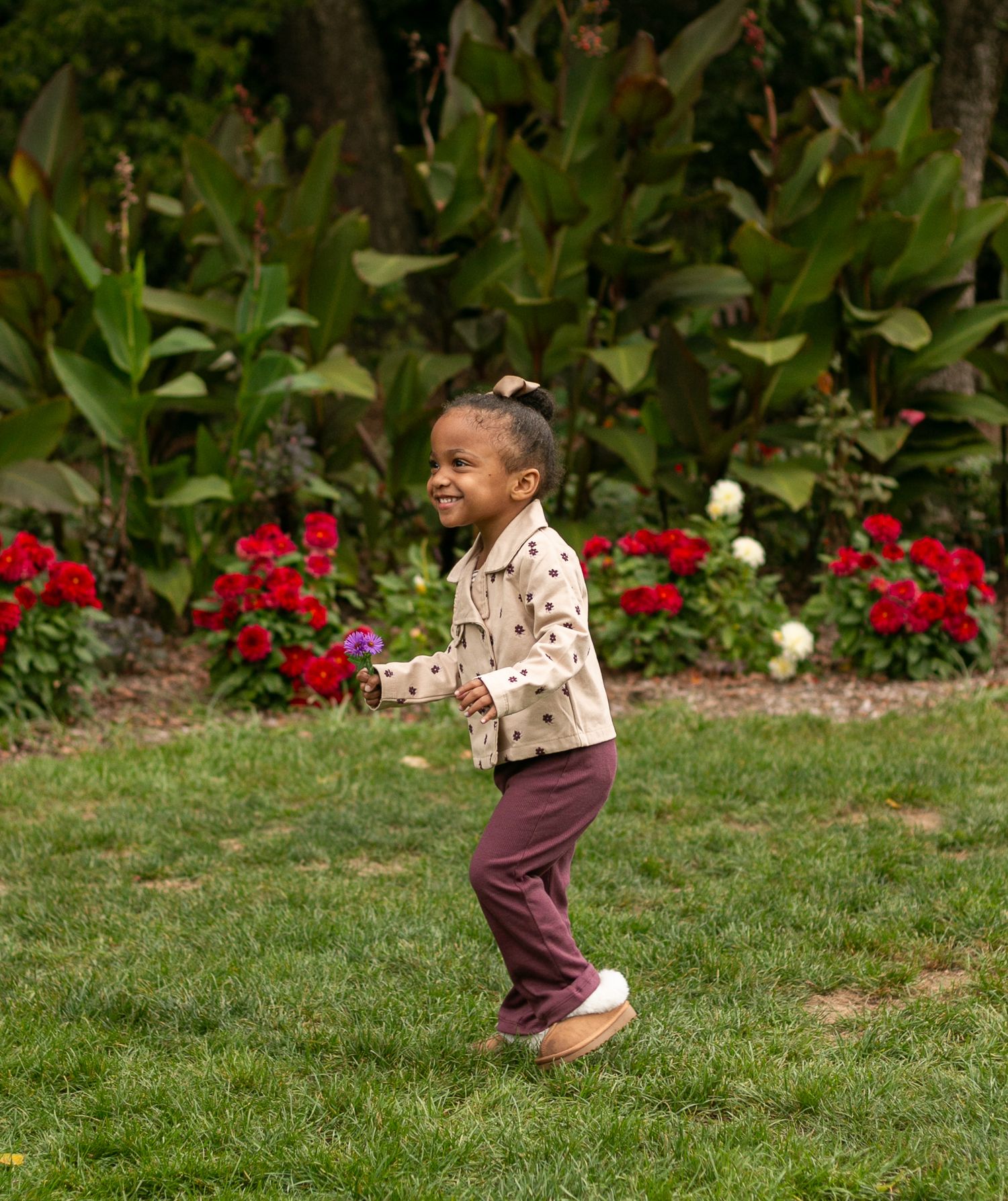 A small child plays and runs joyfully on green grass with vibrant red flowers in the background.