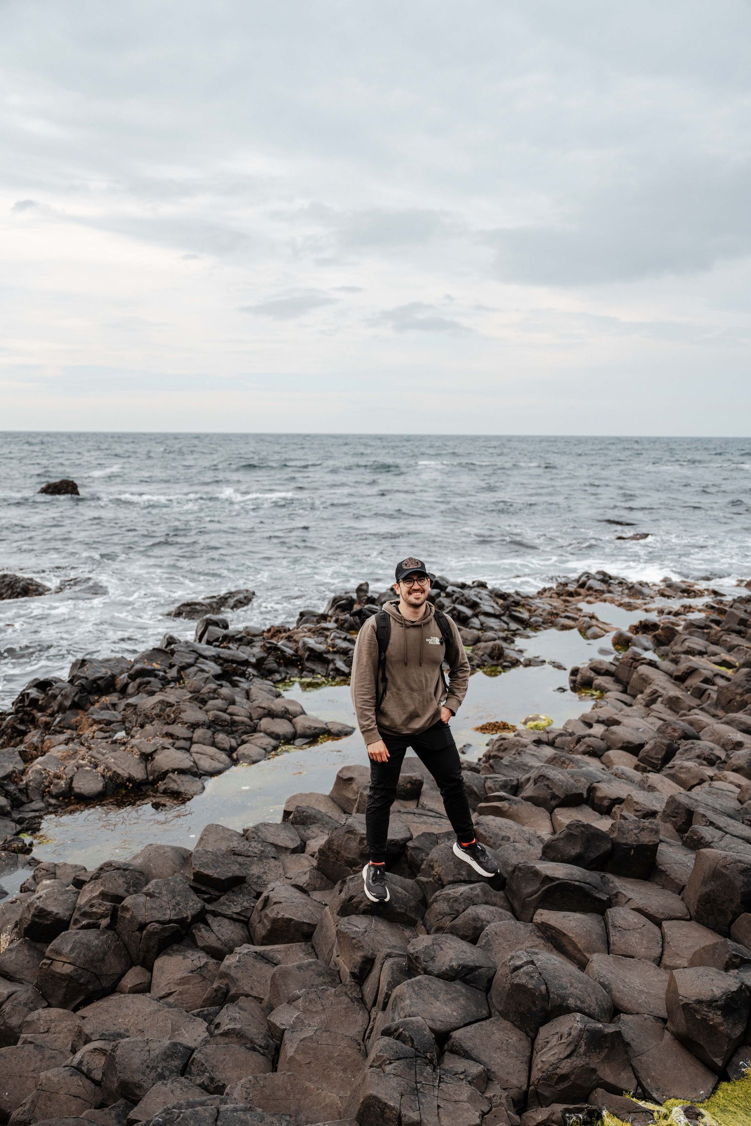 Man standing on rocky coastal shoreline with ocean waves and cloudy sky in the background.