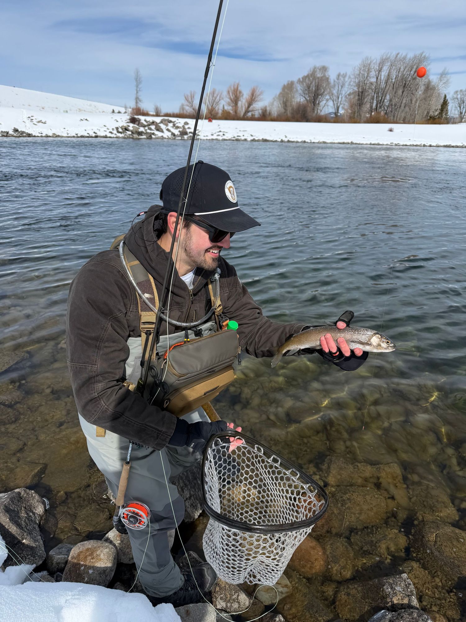 Angler holding a small trout while fly fishing in a winter river with snow on the banks.