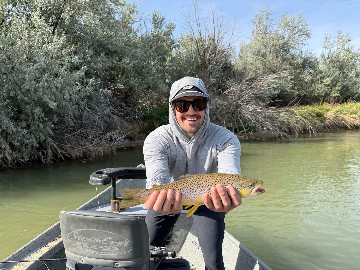 Man in sunglasses holding a brown trout on a fishing boat on a calm river surrounded by green shrubs.