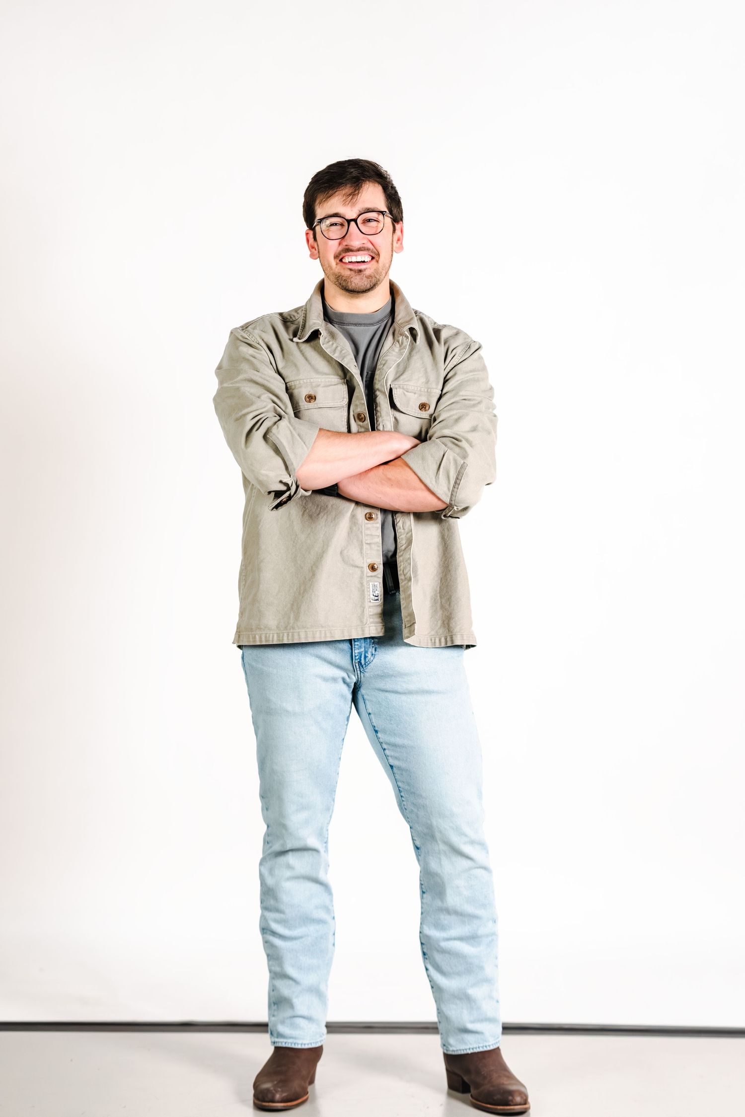 Smiling man with glasses in olive jacket and jeans posing with arms crossed on white background.