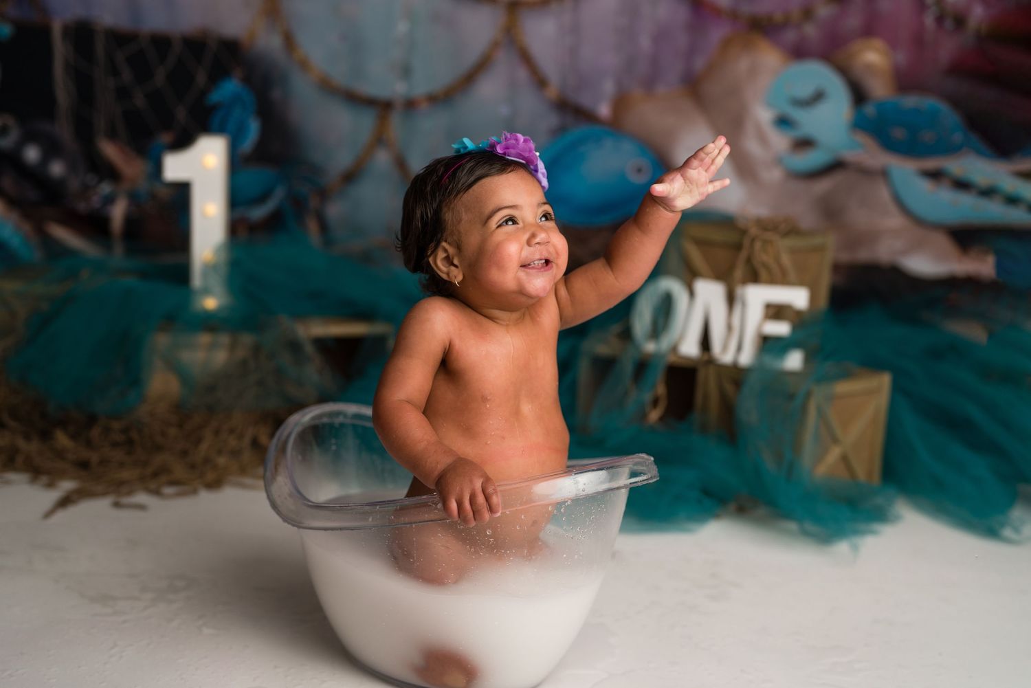Baby in white bowl wearing crown during cake smash photoshoot with number one decoration in background.