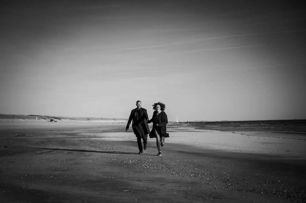 Paar rennt lachend am einsamen Strand auf die Kamera zu, Schwarz-Weiß-Outdoor-Pärchenshooting in Dänemark