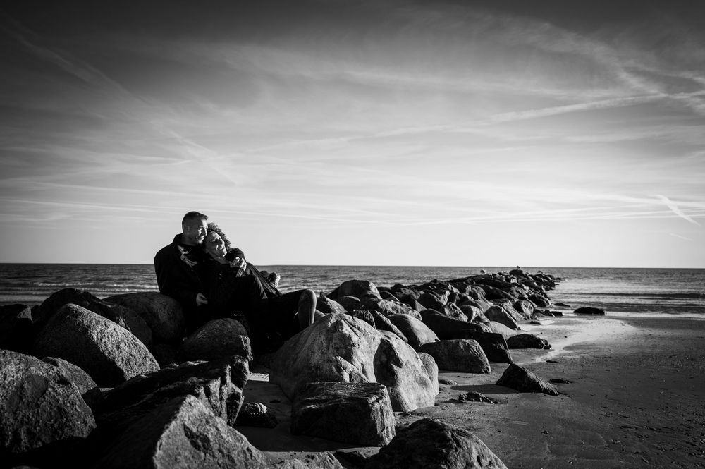 Paar sitzt eng umschlungen auf einem Felsen am Strand, schwarz-weiß Outdoor-Foto