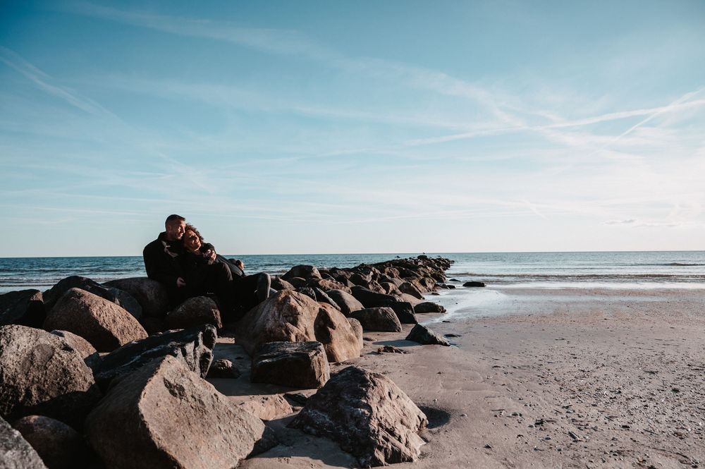 Paar sitzt entspannt auf Stein am Strand, lacht und genießt den Moment, farbiges Pärchenshooting.