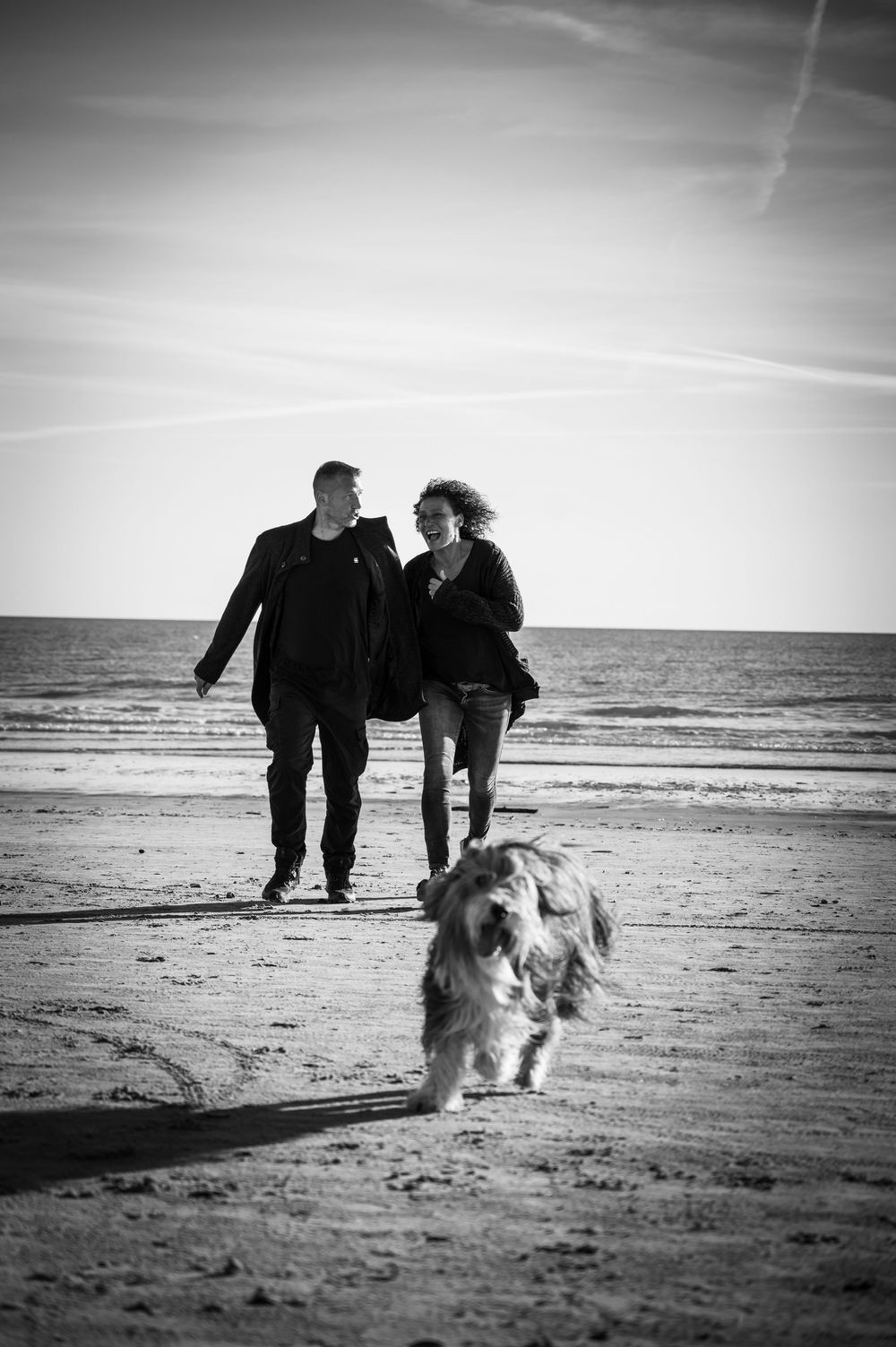 Schwarz-Weiß: Paar wirbelt im Sand umher, Hund mittendrin, authentischer Moment