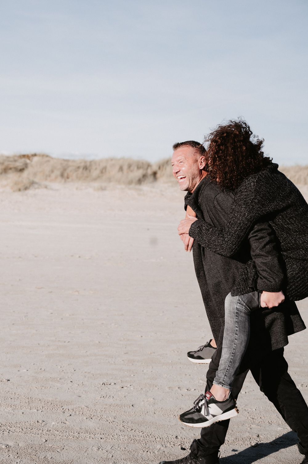 Frau sitzt lachend Huckepack auf den Schultern ihres Partners am Strand in Dänemark, farbiges Outdoor-Pärchenshooting