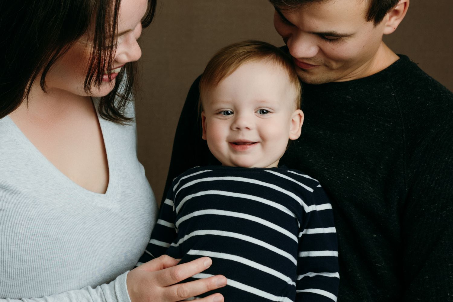 A photo of smiling baby wearing a black and white striped shirt is held lovingly between two parents at the family studio in Victoria BC.