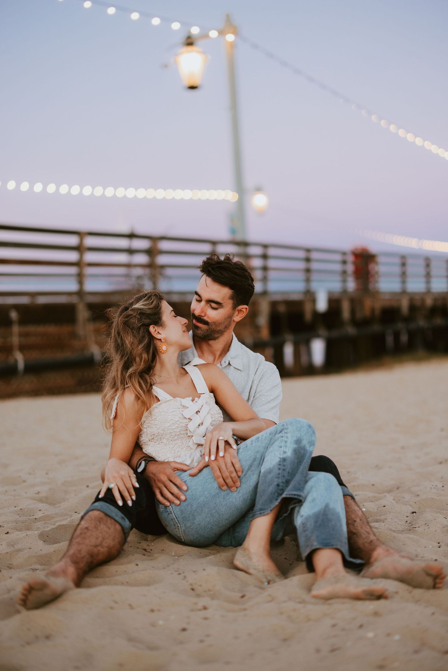 Couple sits closely on a sandy beach at dusk beneath warm string lights near a pier.