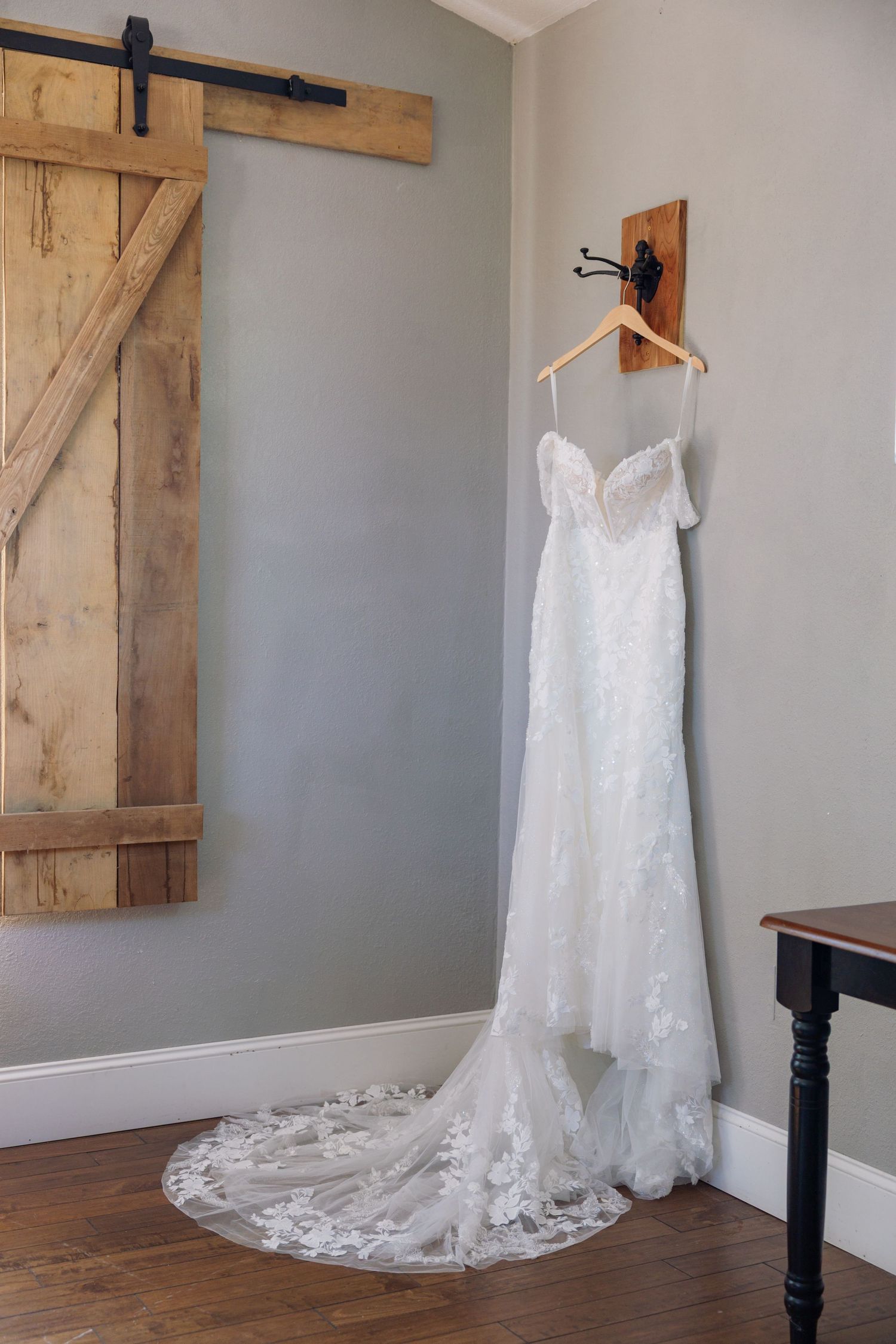 A white lace wedding dress hangs elegantly against a gray wall with rustic barn door decor in a bridal suite.