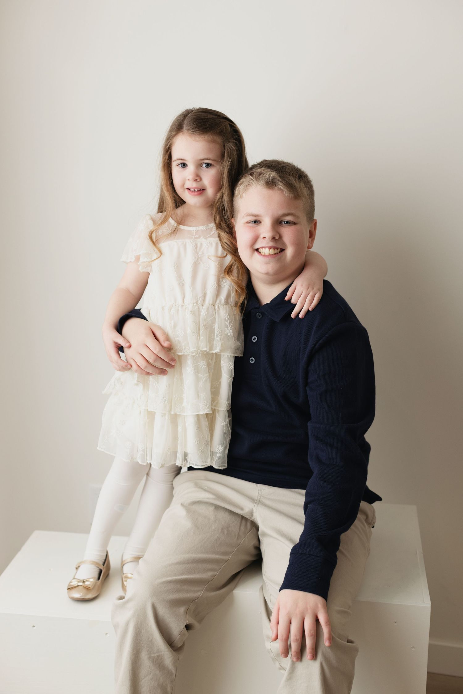 Two children in formal attire pose together against a neutral backdrop in a series of sweet sibling portraits.