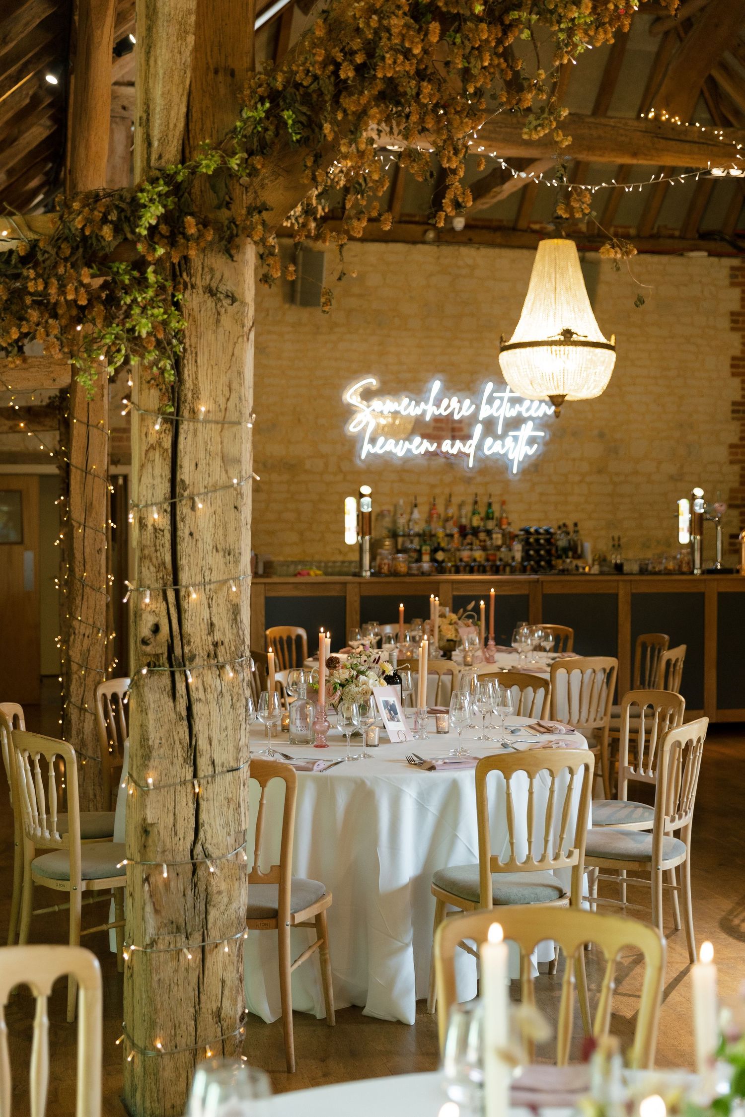 Rustic wedding reception venue with white tablecloths, gold chairs, twinkle lights and a neon sign on exposed brick wall.