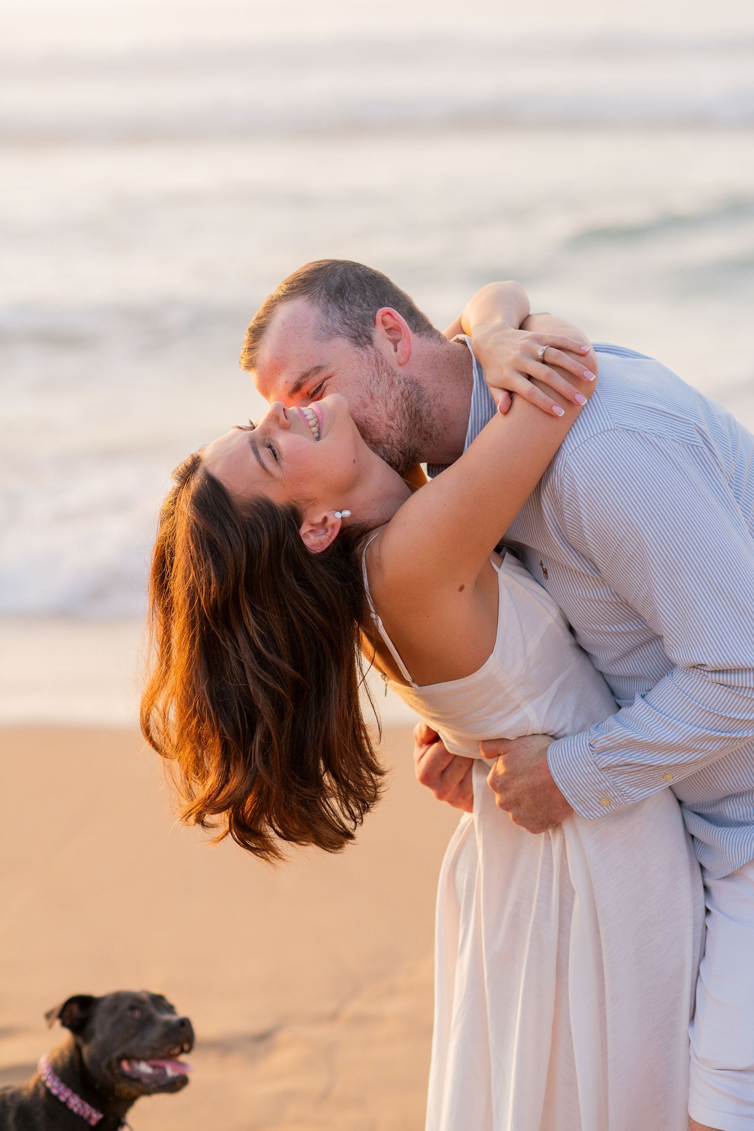 A romantic couple shares a passionate kiss on the beach at sunset while their dog watches in the background.