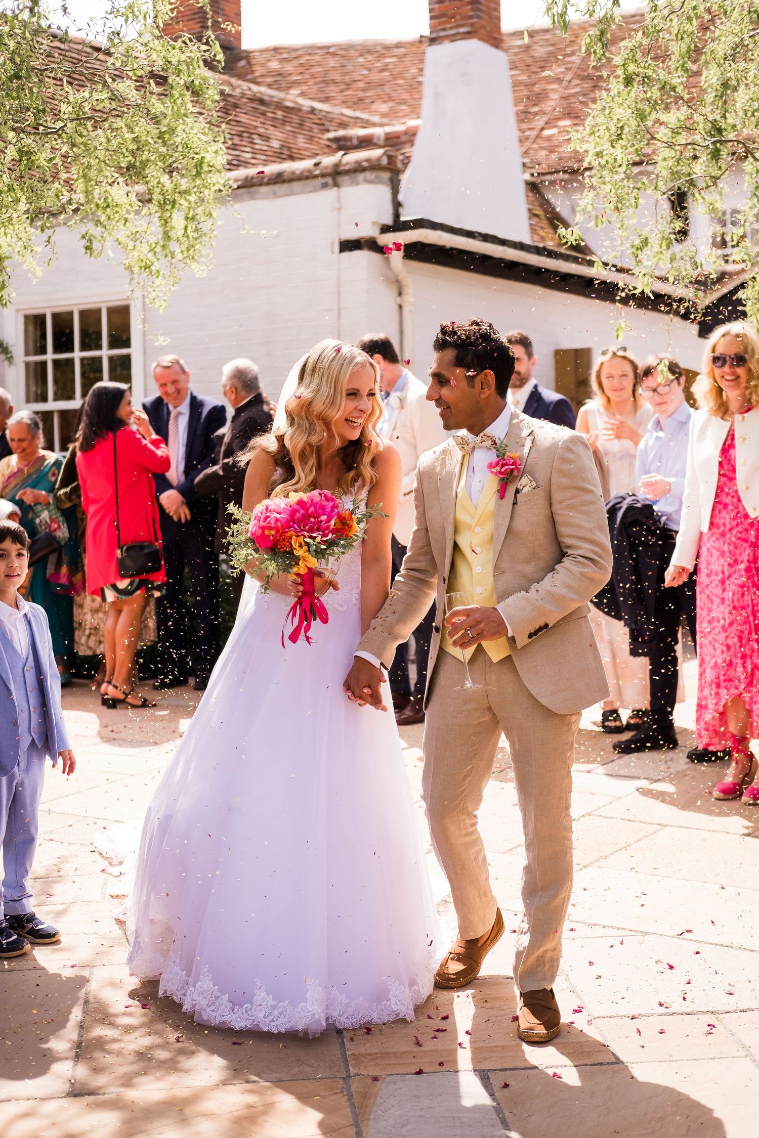 A wedding couple walks together on a sunny patio while guests shower them with confetti, the bride holding vibrant pink flowers.