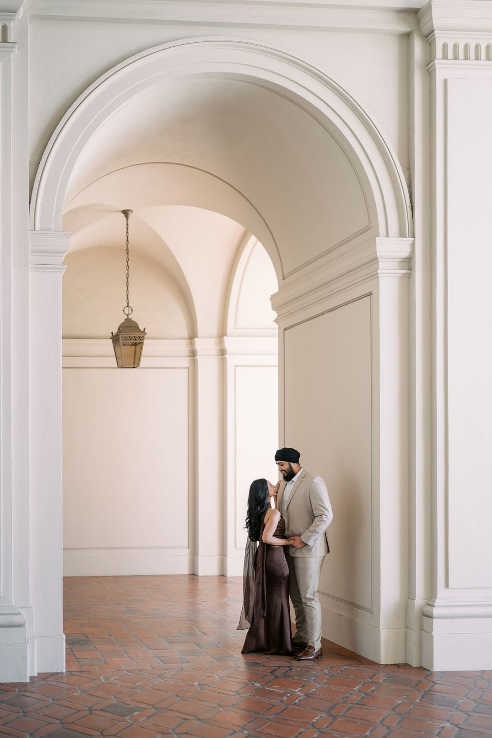 A couple shares an intimate moment under a white arched hallway with terracotta floors and hanging lantern.