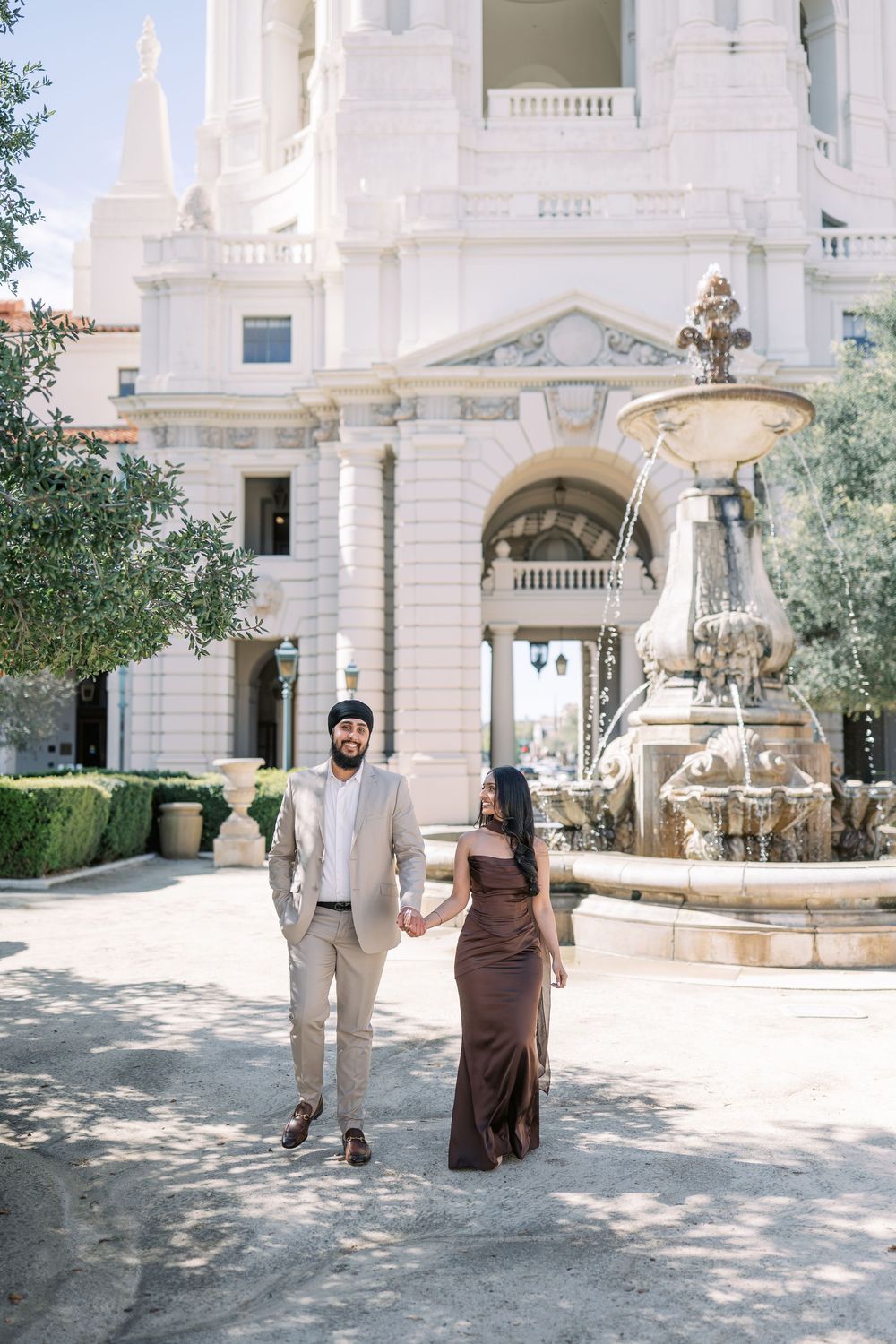 Two people stand in front of an elegant white mansion with ornate architecture and a decorative fountain.