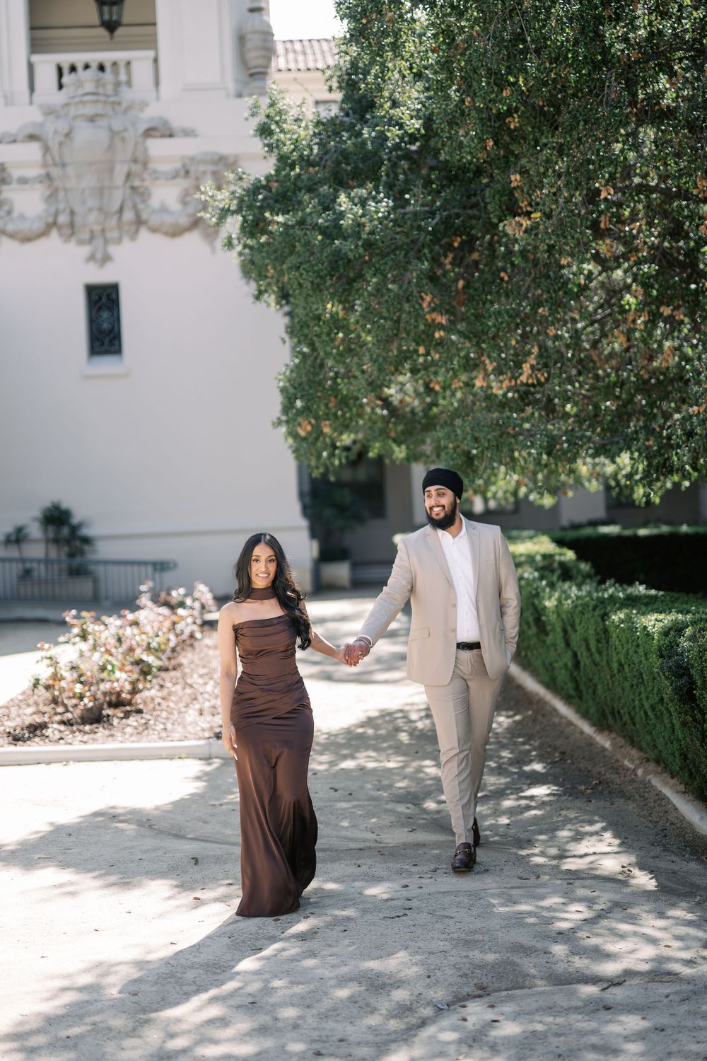 A couple walks and poses together in an outdoor courtyard with trees and brick pathways.