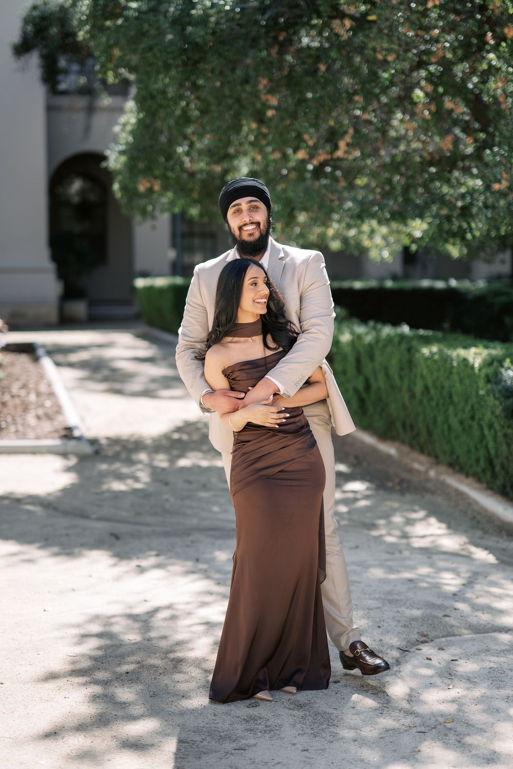 A couple embraces outdoors in formal attire on a sunlit garden path with trees and greenery in the background.