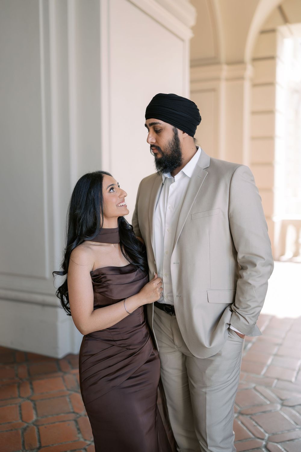 A couple shares intimate moments in formal attire against a light-colored architectural backdrop.