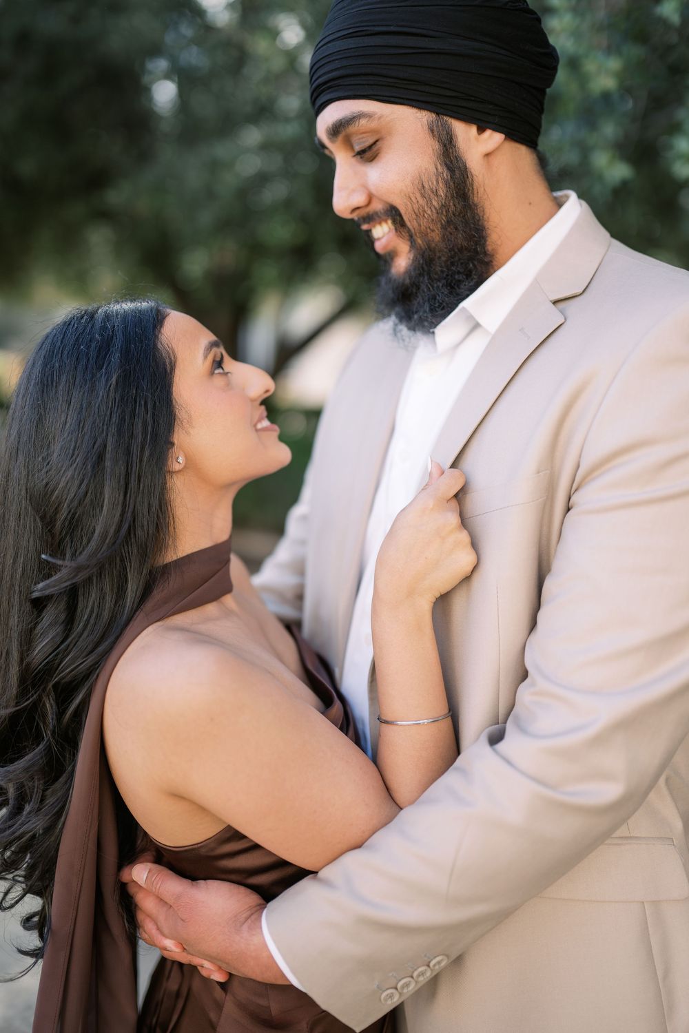 A romantic couple embraces outdoors, with one wearing a beige suit and black turban while the other wears a brown dress.