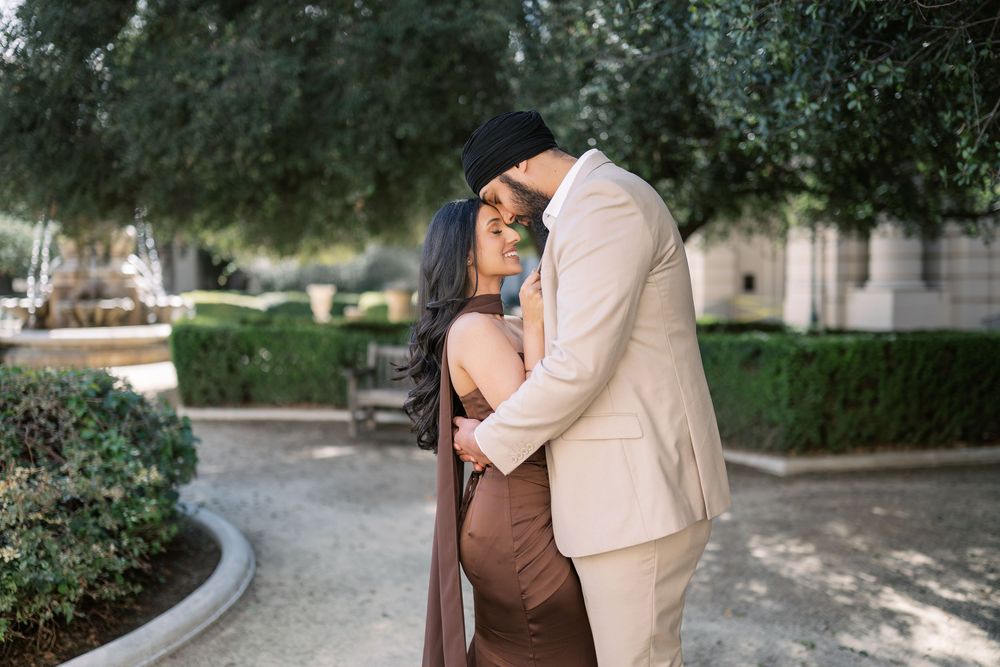 A romantic couple shares a tender moment outdoors in a garden setting wearing coordinating beige and brown outfits.