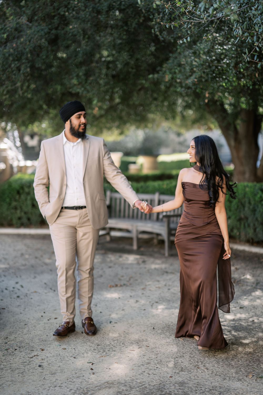 A couple in coordinated beige and brown formal attire pose together in a romantic outdoor garden setting.