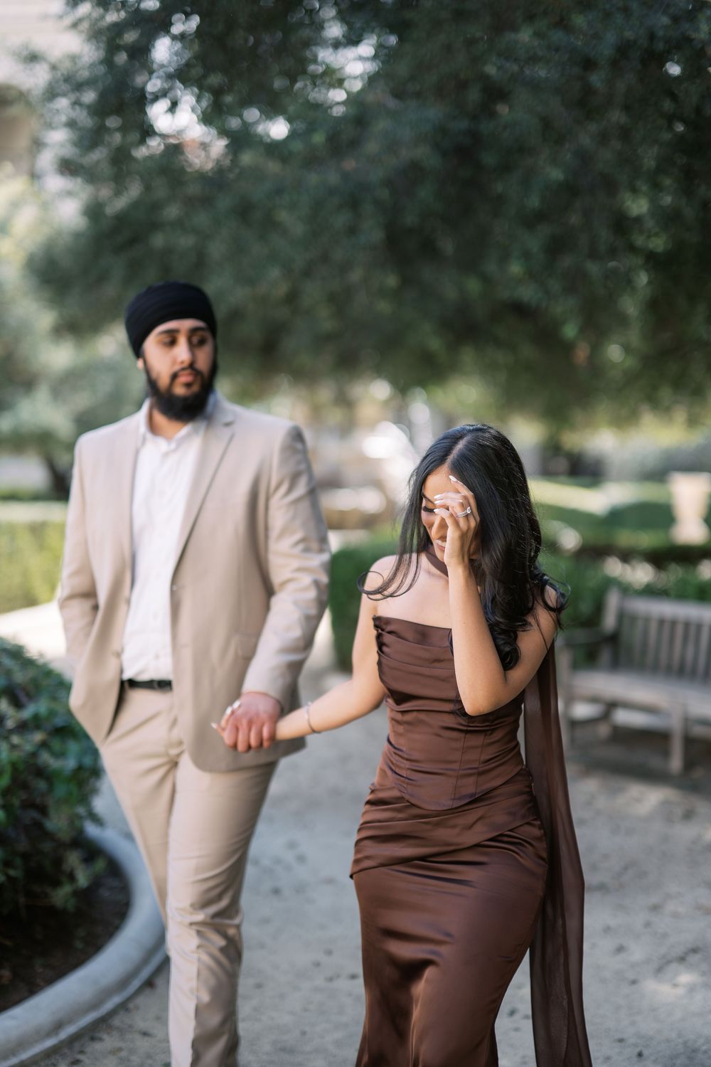 A couple in brown dress and beige suit poses playfully in a park setting during a photo session.