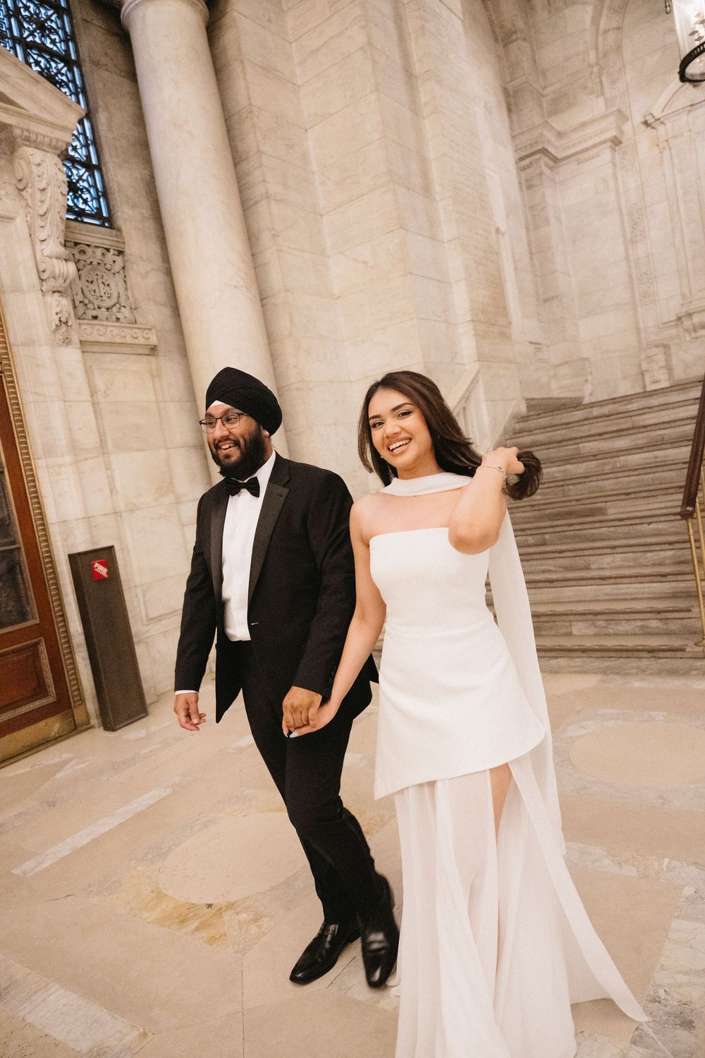 Sikh groom in black tuxedo and turban walks with bride in white strapless gown at elegant stone building.