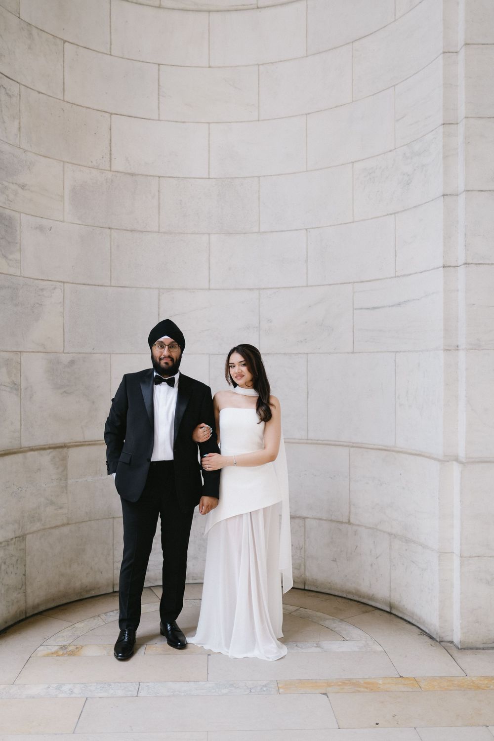 Sikh groom in black tuxedo and turban poses with bride in white gown against marble wall.
