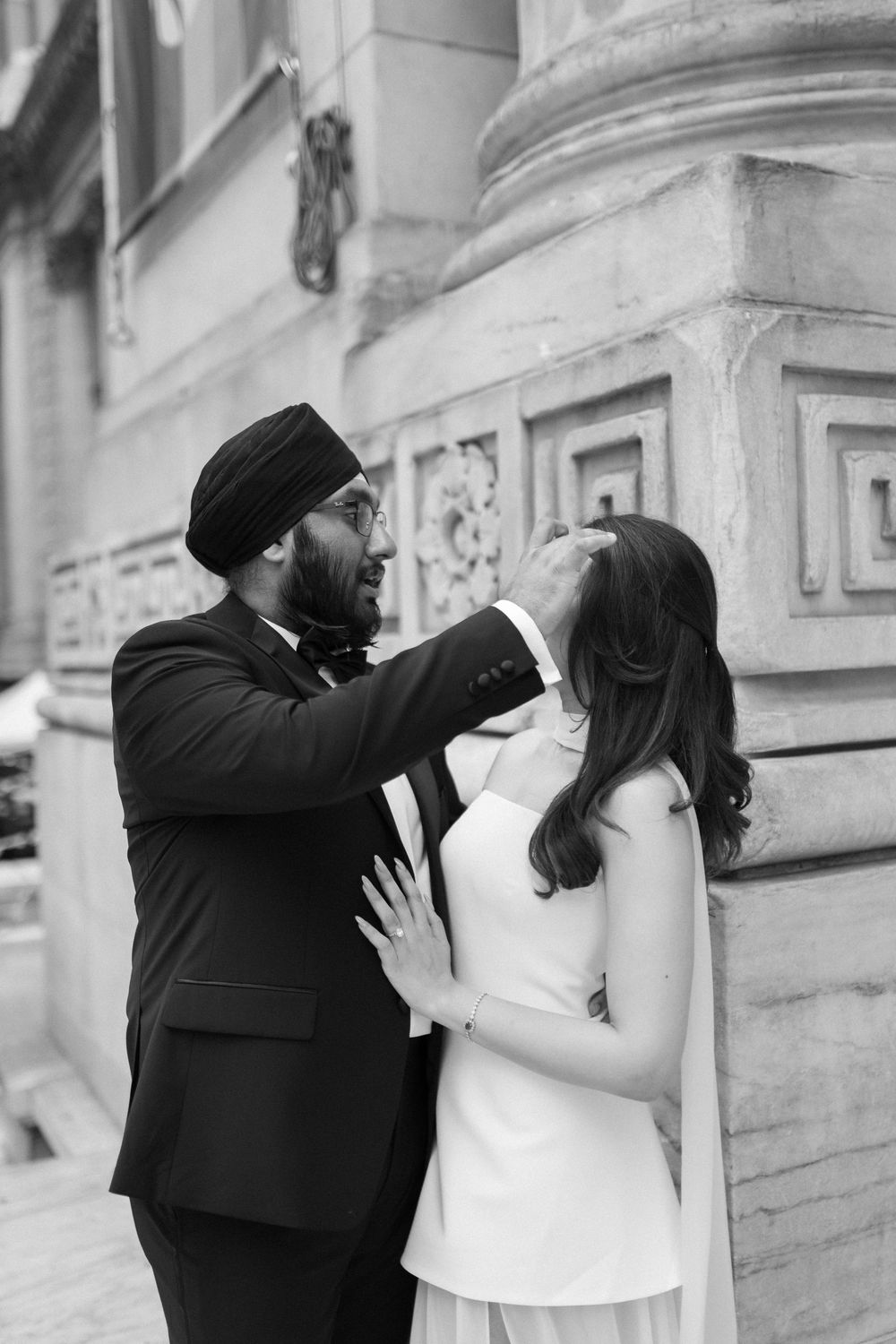 Black and white photo of Sikh groom tenderly touching brides face outside ornate stone building.