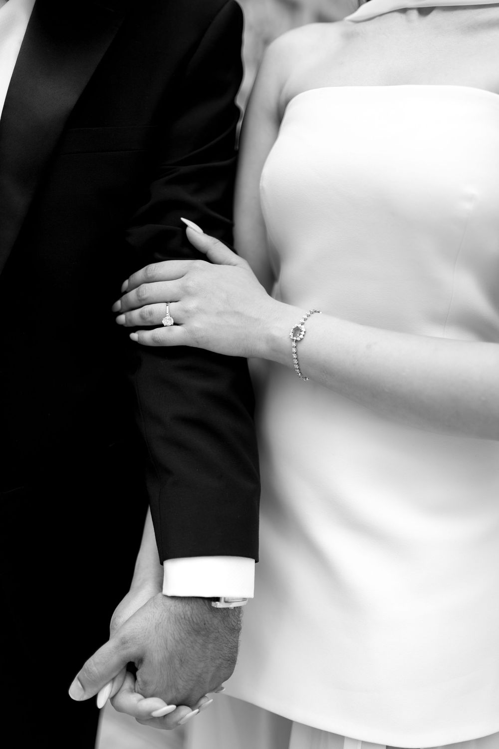 Black and white photo of bride and groom holding hands, showcasing engagement ring and bracelet.