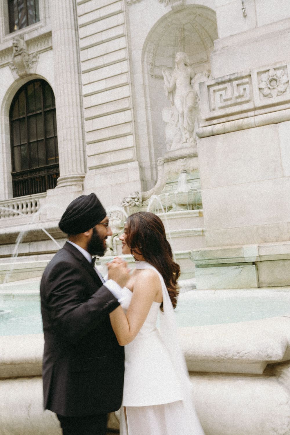 Sikh groom in black tuxedo and bride in white gown share a romantic moment by a classical stone fountain.