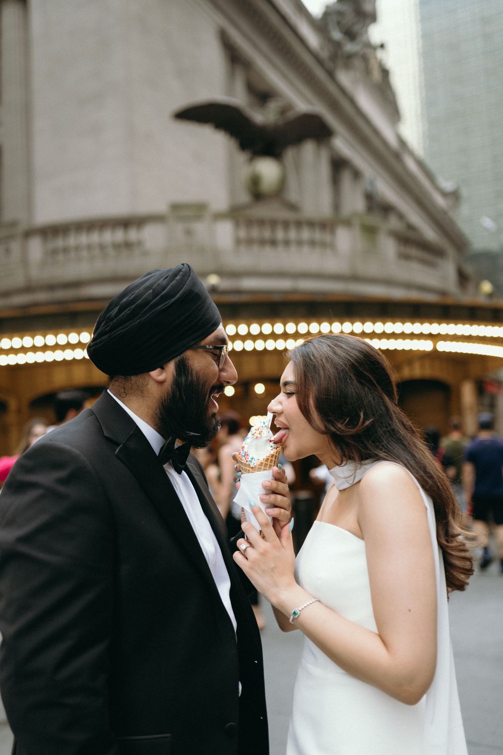 Sikh groom and bride in white dress share ice cream cone outside Grand Central Terminal.