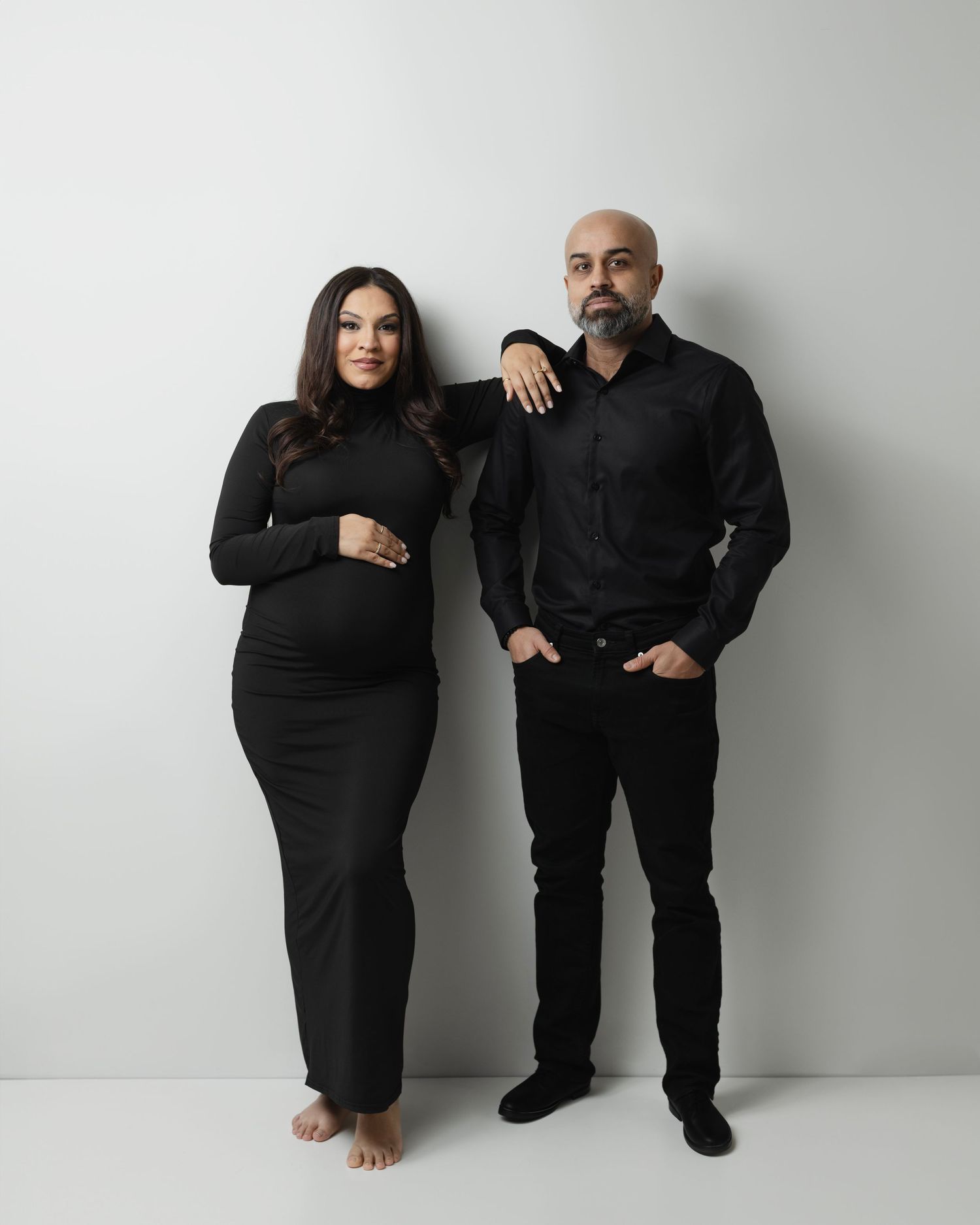A couple in matching black outfits poses together against a white wall in a minimalist studio setting. Elizabeth Lang Photography Surrey