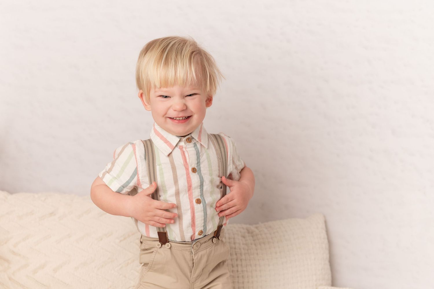 A small child in a striped shirt and khaki shorts sits and plays on a cream-colored backdrop.