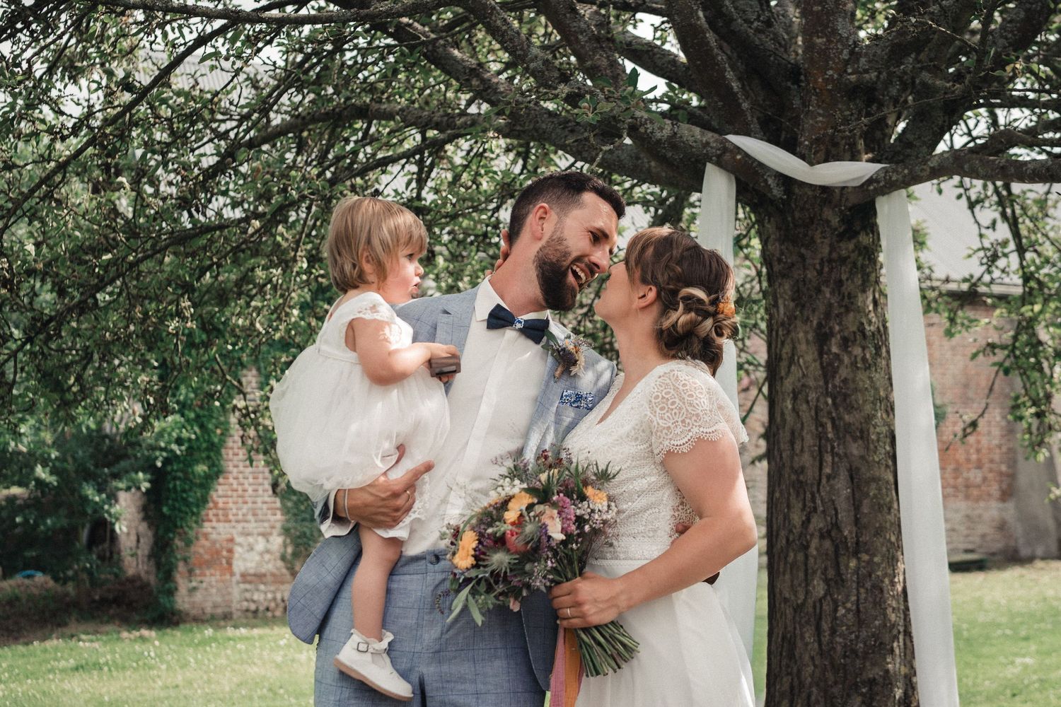 Outdoor wedding ceremony under a large tree with couple and small child holding autumn-inspired floral bouquet.