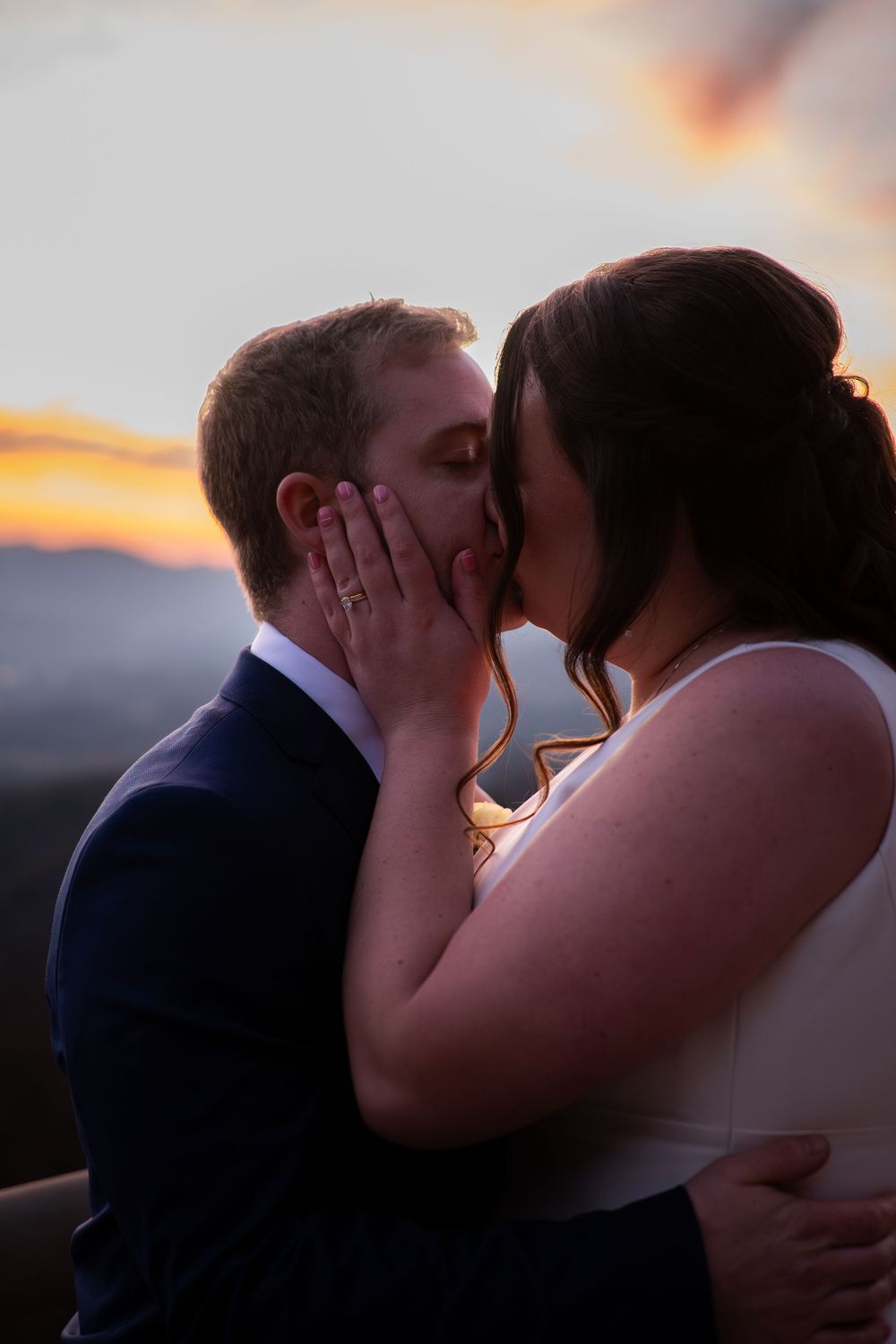 A couple shares a romantic kiss at sunset with mountains in the background.