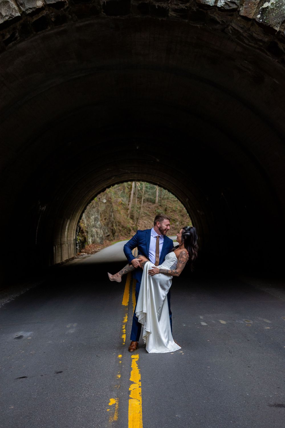 A romantic couple embraces in a dramatic dip pose under a dark tunnel with yellow road lines visible below.