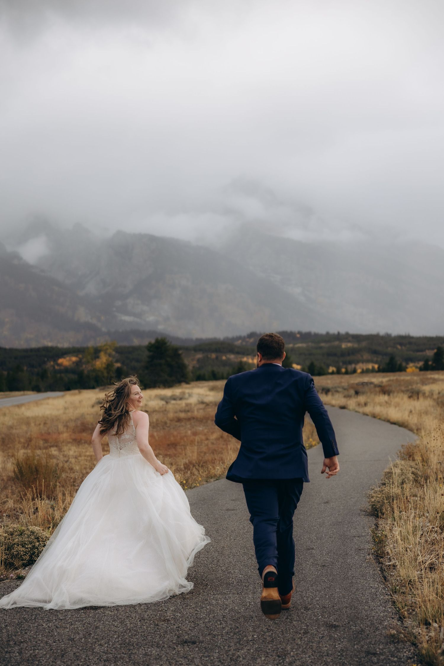 A couple in wedding attire runs playfully down a mountain path on a misty, dramatic day.