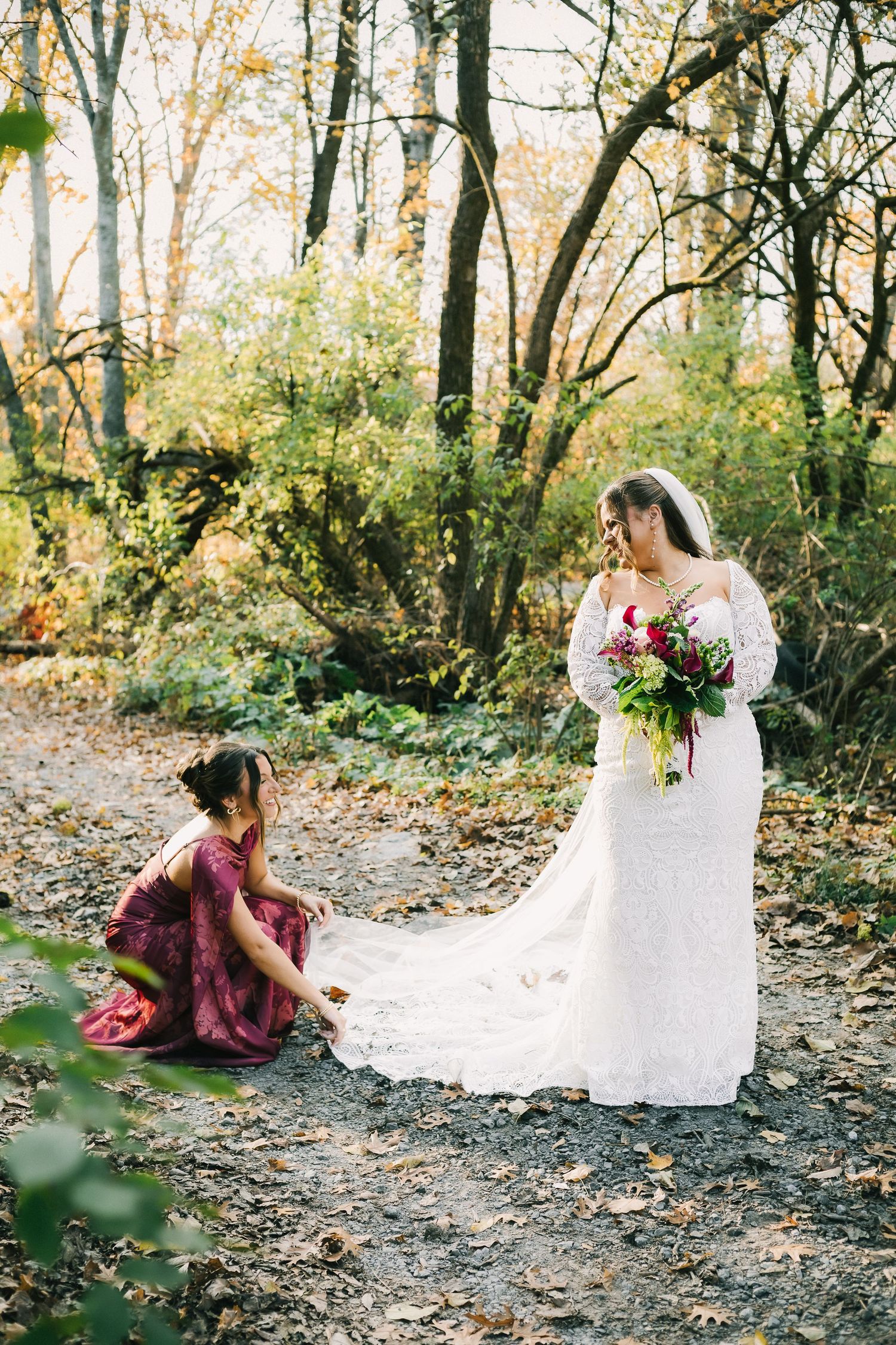 A bride in a flowing white dress sits gracefully on a leaf-covered forest path during an autumn photoshoot.