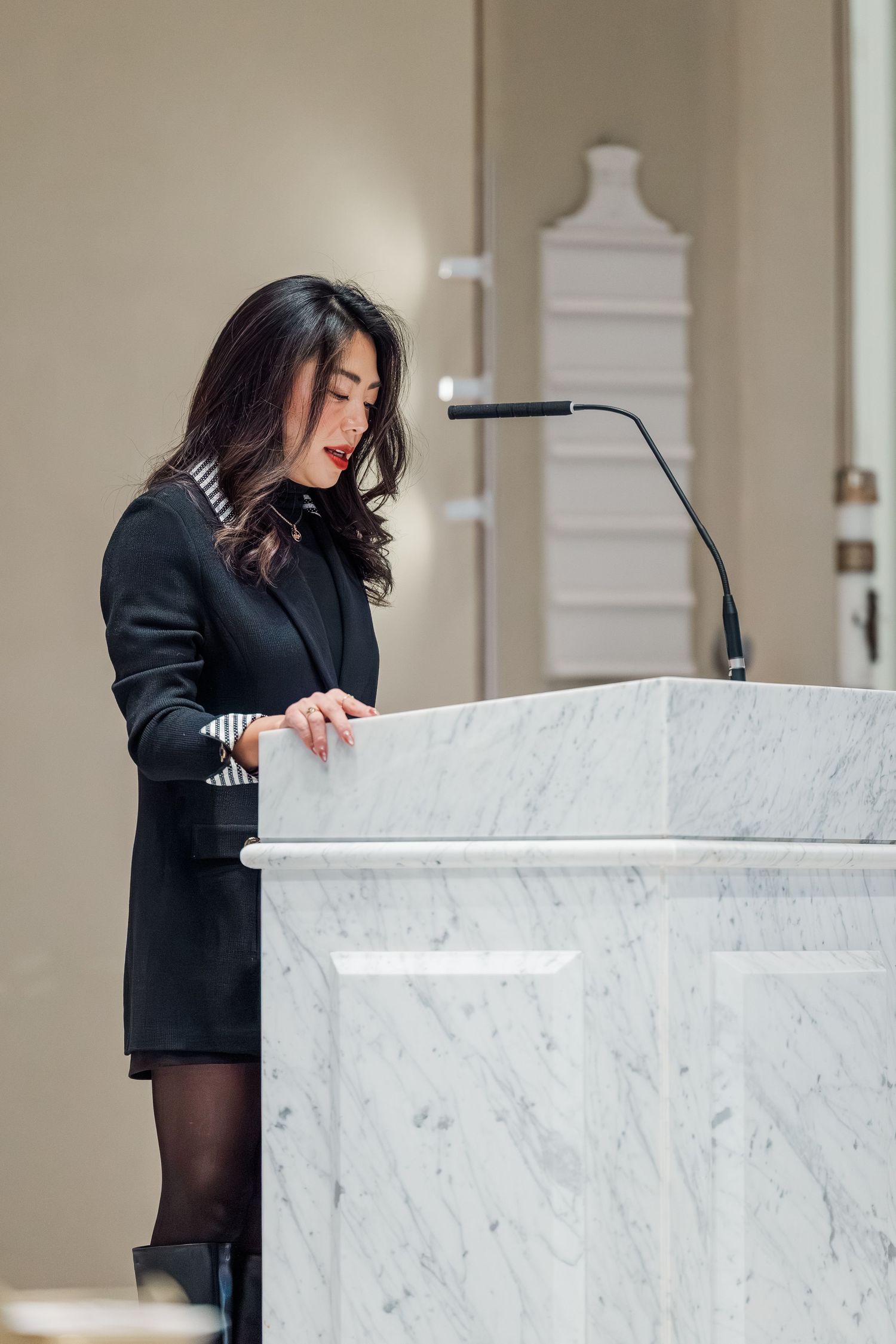 A speaker stands at a white marble podium with a black desk lamp in an elegant indoor setting.