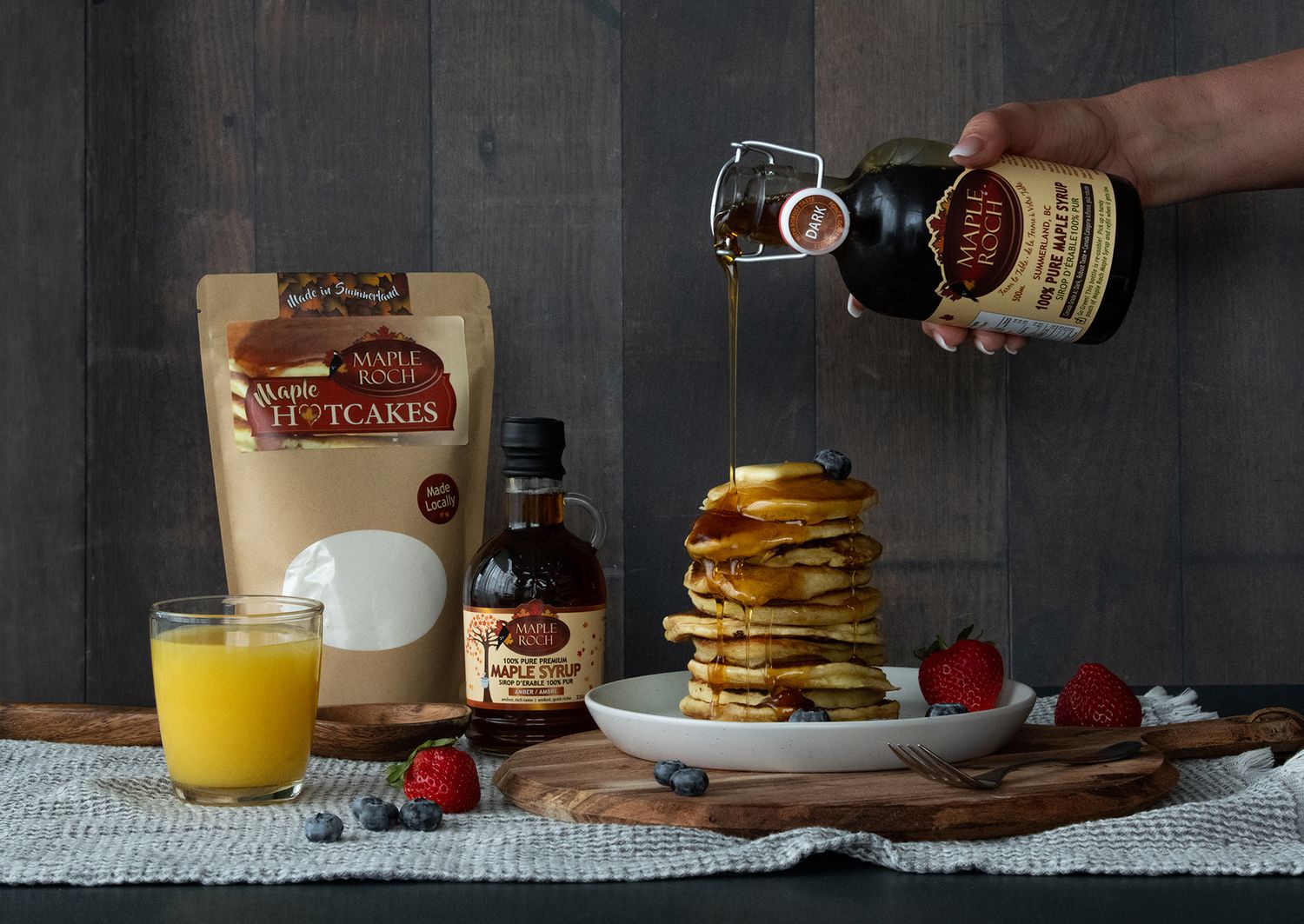 Styled food photography of maple syrup being poured over pancakes with fresh berries and orange juice.