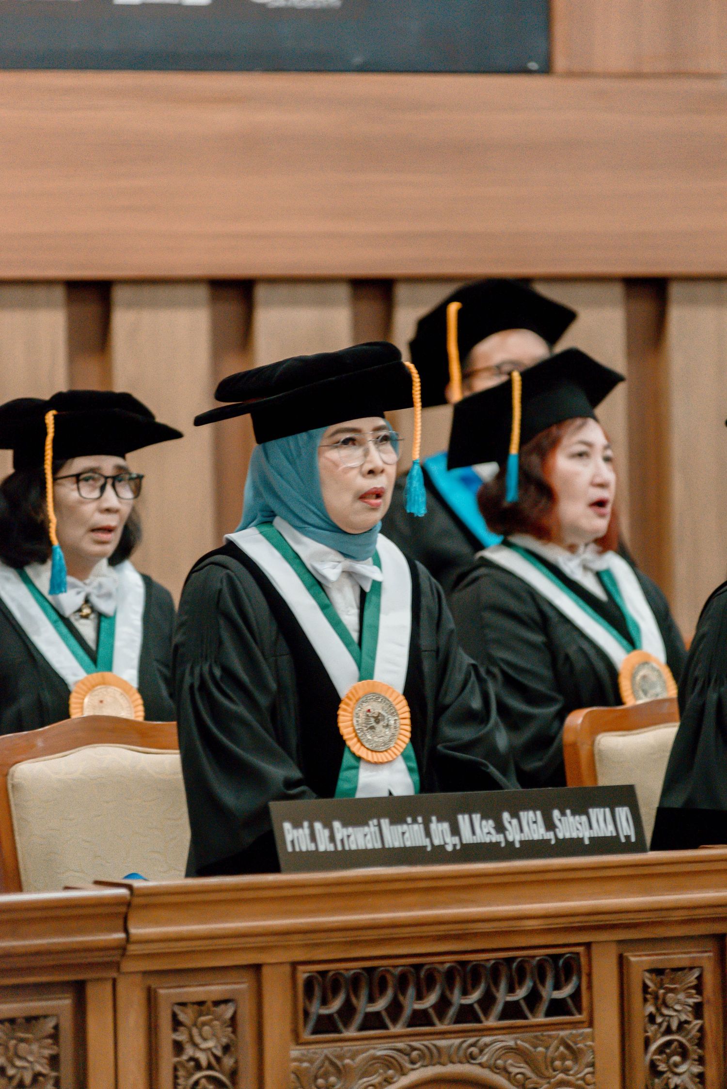 Academic faculty members in graduation regalia seated at a formal ceremony wearing black caps and green stoles.