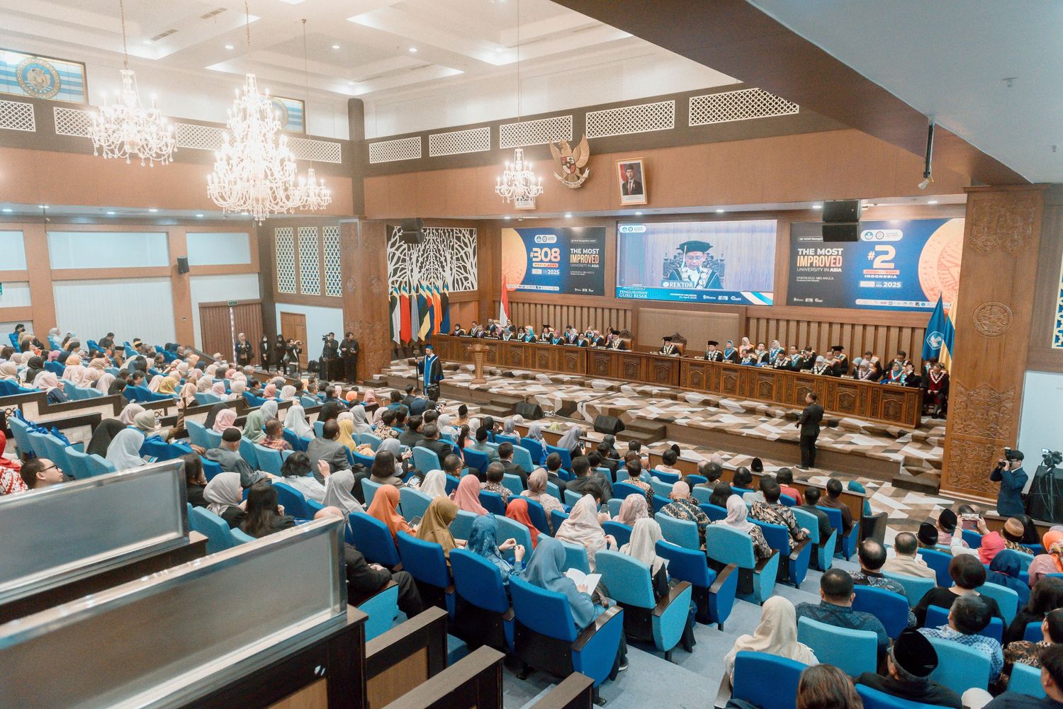 Large auditorium filled with attendees in blue uniforms during a formal ceremony with flags and video screens visible.