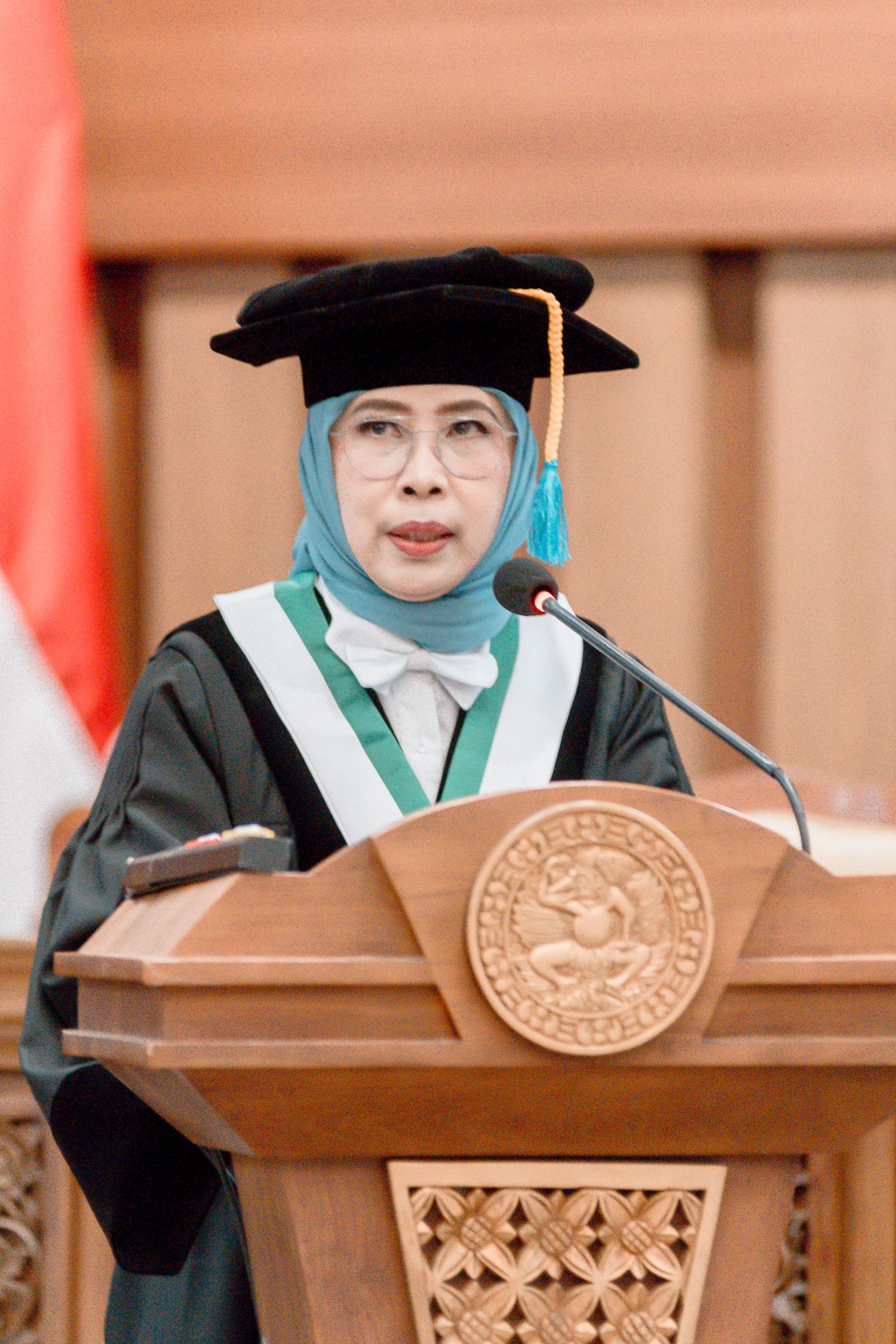 Graduate wearing academic regalia and graduation cap stands at wooden podium during commencement ceremony.
