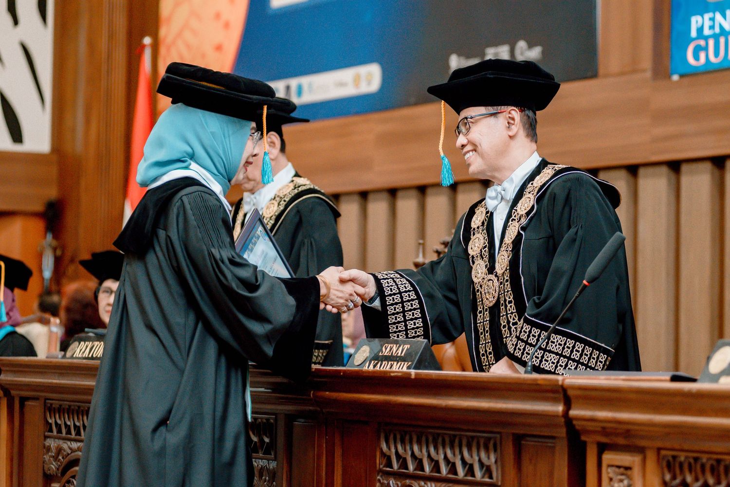 Two people in academic regalia shake hands during a graduation ceremony in a wood-paneled hall.