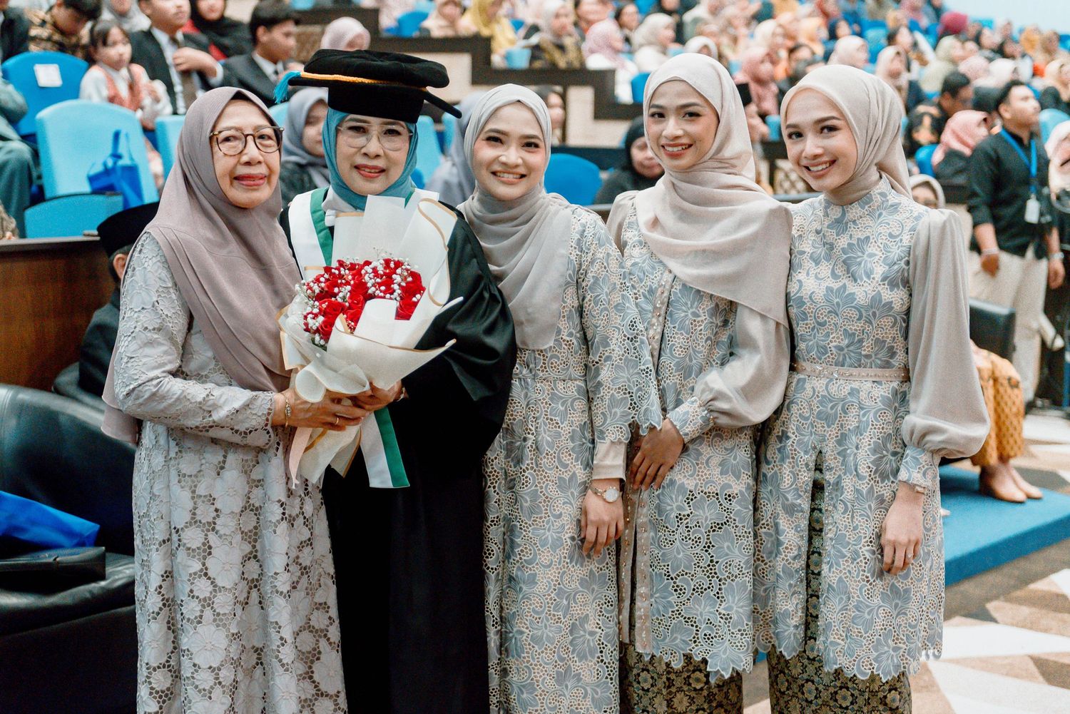 Group of women in matching floral lace dresses and hijabs pose together at an outdoor celebration.