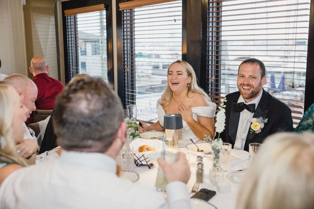 Wedding reception guests seated at elegant tables with white linens near large windows in a restaurant setting.