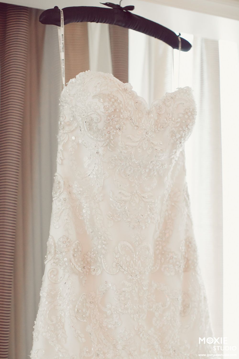 White lace wedding dress hanging in front of a window with soft natural light filtering through.