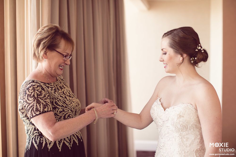 Wedding party helps bride get dressed in elegant hotel room with soft lighting and curtains.